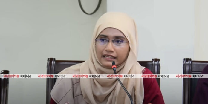 হিজাব পরিহিত সামান্তা শারমিন একটি সংবাদ সম্মেলনে মাইক্রোফোনের সামনে কথা বলছেন এবং অন্তর্বর্তীকালীন সরকারের সমালোচনা করছেন।